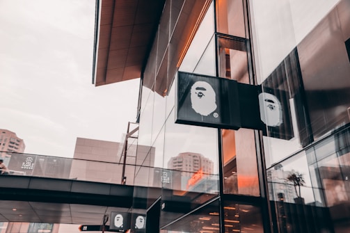 A modern storefront with reflective glass panels and an elevated walkway showing the logo of a brand featuring a stylized ape face. The architecture is sleek with an urban environment reflected in the windows, under a cloudy sky.