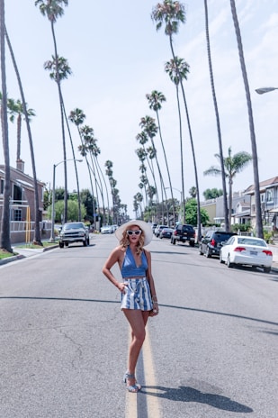 A person wearing a stylish summer outfit, including a sunhat, sunglasses, a blue halter top, and striped shorts, standing confidently in the middle of a palm tree-lined street. The street is flanked by houses and parked cars under a clear blue sky.
