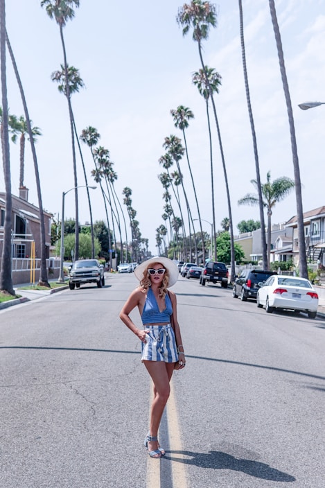 A person wearing a stylish summer outfit, including a sunhat, sunglasses, a blue halter top, and striped shorts, standing confidently in the middle of a palm tree-lined street. The street is flanked by houses and parked cars under a clear blue sky.