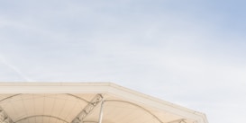 A modern architectural structure featuring a partially visible triangular roof with a beige fabric cover, supported by metal beams. The sky in the background is clear and light blue.