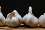 Several heads of garlic are arranged on a wooden surface with a dark background. The garlic bulbs show a natural white outer skin with some brown roots attached.