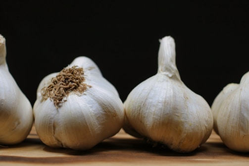 Several heads of garlic are arranged on a wooden surface with a dark background. The garlic bulbs show a natural white outer skin with some brown roots attached.