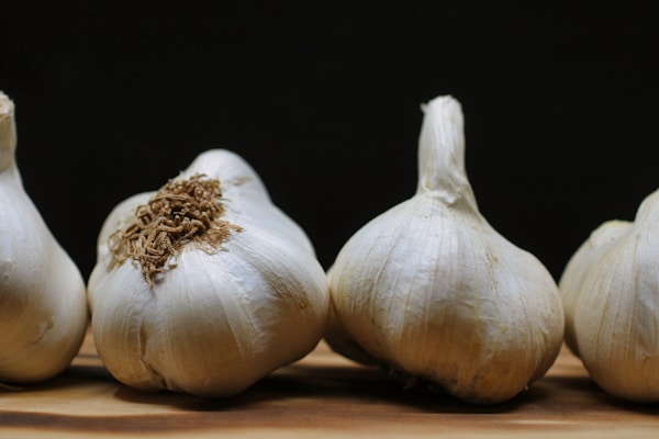 Several heads of garlic are arranged on a wooden surface with a dark background. The garlic bulbs show a natural white outer skin with some brown roots attached.