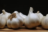 Several heads of garlic are placed on a wooden surface. The garlic heads feature off-white papery skins, with one in the foreground showing some dried roots. The background is dark, creating a strong contrast with the garlic.