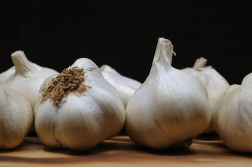 Several heads of garlic are placed on a wooden surface. The garlic heads feature off-white papery skins, with one in the foreground showing some dried roots. The background is dark, creating a strong contrast with the garlic.