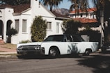 A vintage car is parked on a suburban street in front of a white stucco house with a red tiled roof. Palm trees and mountains are visible in the background, contributing to a serene neighborhood atmosphere.