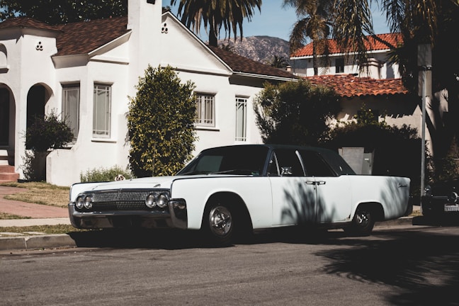 A vintage car is parked on a suburban street in front of a white stucco house with a red tiled roof. Palm trees and mountains are visible in the background, contributing to a serene neighborhood atmosphere.