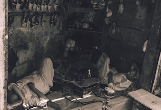 Two men are resting inside a small, dimly lit workshop. They are lying on the concrete floor amidst scattered tools and objects. The shelves above them are filled with pairs of traditional shoes. The setting appears rustic and has an air of simplicity.