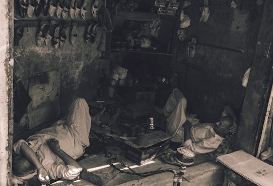 Two men are resting inside a small, dimly lit workshop. They are lying on the concrete floor amidst scattered tools and objects. The shelves above them are filled with pairs of traditional shoes. The setting appears rustic and has an air of simplicity.