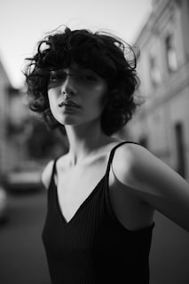 A black and white portrait of a person with curly hair and a nose ring, wearing a sleeveless top, standing outdoors on a paved street with blurred buildings in the background.