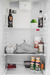 Inside a refrigerator, several food and drink items are organized on the shelves. On the top shelf, there's a bottle of Guinness beer, a Voss water bottle, a black bottled drink, and a small potted succulent plant. The middle shelf unexpectedly contains a single grey sneaker, a carton of eggs, a bottle of milk, and jars of condiments. The bottom shelf includes packs of cheese or similar items and two bottles of Bio Chefir yogurt. The refrigerator interior is bright and clean.