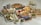 A variety of foods are arranged on a table, including pizzas, tacos, a fresh salad, bowls of salsa and guacamole, tortilla chips, and fresh fruit like apples and avocados. Metal salt and pepper shakers, as well as bottles that appear to contain sauces, are also present.
