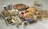 A variety of foods are arranged on a table, including pizzas, tacos, a fresh salad, bowls of salsa and guacamole, tortilla chips, and fresh fruit like apples and avocados. Metal salt and pepper shakers, as well as bottles that appear to contain sauces, are also present.