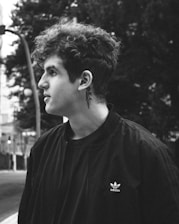 A black and white portrait of a person with curly hair and a visible neck tattoo, wearing a dark Adidas jacket. The person is looking to the side, and the background is an urban setting with trees and out-of-focus streetlights.