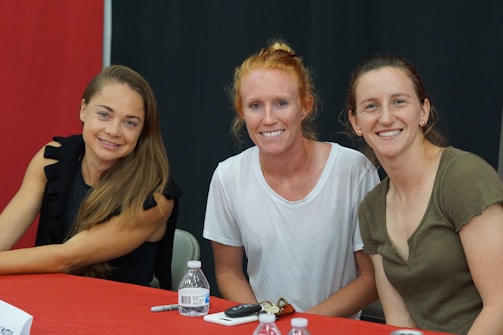 Three women are seated at a table with a red and black backdrop. They are smiling at the camera, with two of them wearing casual tops and the third in a black, ruffled sleeve top. A water bottle, marker, and some papers are placed on the table.