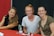 Three women are seated at a table with a red and black backdrop. They are smiling at the camera, with two of them wearing casual tops and the third in a black, ruffled sleeve top. A water bottle, marker, and some papers are placed on the table.