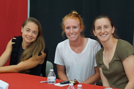 Three women are seated at a table with a red and black backdrop. They are smiling at the camera, with two of them wearing casual tops and the third in a black, ruffled sleeve top. A water bottle, marker, and some papers are placed on the table.