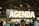 A large sign with the word 'Agenda' is prominently displayed, illuminated with bright lights. The setting appears to be an indoor event or trade show, with numerous people gathered below the sign. The ceiling features industrial elements such as pipes and large lights.