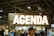 A large sign with the word 'Agenda' is prominently displayed, illuminated with bright lights. The setting appears to be an indoor event or trade show, with numerous people gathered below the sign. The ceiling features industrial elements such as pipes and large lights.