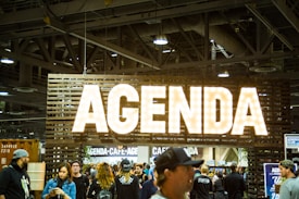 A large sign with the word 'Agenda' is prominently displayed, illuminated with bright lights. The setting appears to be an indoor event or trade show, with numerous people gathered below the sign. The ceiling features industrial elements such as pipes and large lights.