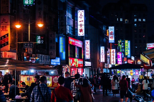A bustling night market scene with numerous brightly illuminated signs in various colors and Chinese characters. Many people are walking along the street, some are browsing stalls while others sit at makeshift tables enjoying food. Shops and food vendors line the streets, creating a vibrant and lively atmosphere.