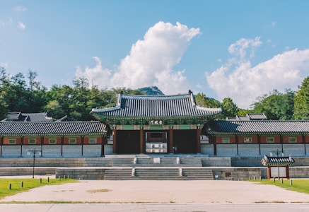 A traditional Asian-style building with ornate architectural details, featuring a symmetrical design and intricate roof patterns. The scene is set in a tranquil environment with lush green trees in the background and a dramatic blue sky filled with white clouds.
