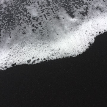 White sea foam spreads over dark sand, creating a striking contrast between the two textures. The foam appears frothy and dynamic, with a distinct separation line where it meets the smooth, black surface.