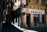 A narrow street with shadows obscuring parts of the scene. On the right, a retro-styled shop with a sign reading 'SERIE B' is visible. The shop has a weathered facade with two white chairs placed outside near a small table. Overhead, there are traditional balconies with ornate railings, and the buildings have various textures and colors. A round traffic sign is visible next to the shop. A car is parked on the left side of the street, adding to the urban feel.