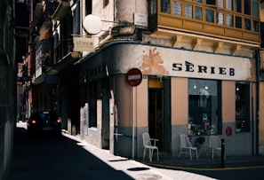 A narrow street with shadows obscuring parts of the scene. On the right, a retro-styled shop with a sign reading 'SERIE B' is visible. The shop has a weathered facade with two white chairs placed outside near a small table. Overhead, there are traditional balconies with ornate railings, and the buildings have various textures and colors. A round traffic sign is visible next to the shop. A car is parked on the left side of the street, adding to the urban feel.
