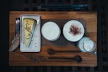 A slice of cheesecake garnished with herbs and crumbs is placed on a rectangular white plate next to a folded napkin with a fork and knife. Two cups filled with frothy beverages sit on a wooden board, one of the cups is dusted with cocoa powder. A small pitcher containing a creamy liquid is beside the cups.