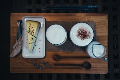 A slice of cheesecake garnished with herbs and crumbs is placed on a rectangular white plate next to a folded napkin with a fork and knife. Two cups filled with frothy beverages sit on a wooden board, one of the cups is dusted with cocoa powder. A small pitcher containing a creamy liquid is beside the cups.