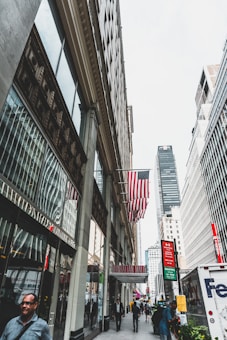 A bustling city street lined with tall buildings. The American flag is prominently displayed, and people are walking along the sidewalk. Signs for shops and businesses, including a 'Manhattan Mall' entrance, are visible. The atmosphere is busy and urban, with vehicles and pedestrians navigating the space.