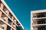 Two modern apartment buildings with multiple floors and balconies stand against a clear blue sky. The buildings have a minimalist design with clean lines, featuring large glass windows and neutral colors.