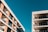Two modern apartment buildings with multiple floors and balconies stand against a clear blue sky. The buildings have a minimalist design with clean lines, featuring large glass windows and neutral colors.