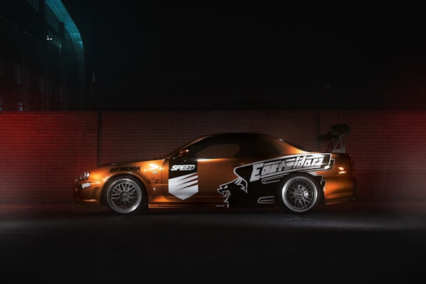 A modified sports car is parked against a brick wall, illuminated by dim lighting. The car features custom decals and a large rear spoiler, with alloy wheels visible. The setting appears to be an urban environment, with a dark, moody atmosphere.