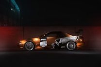 A modified sports car is parked against a brick wall, illuminated by dim lighting. The car features custom decals and a large rear spoiler, with alloy wheels visible. The setting appears to be an urban environment, with a dark, moody atmosphere.