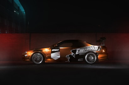 A modified sports car is parked against a brick wall, illuminated by dim lighting. The car features custom decals and a large rear spoiler, with alloy wheels visible. The setting appears to be an urban environment, with a dark, moody atmosphere.