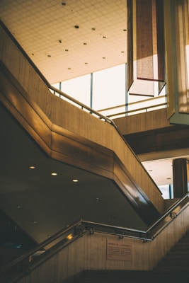 An interior view featuring a modern staircase made of wood and metal with a geometric design. The ceiling is composed of a grid of small lights, giving it a structured appearance. Tall, rectangular window panels allow natural light to enter the space, and several elongated, vertical decor pieces hang from the ceiling.