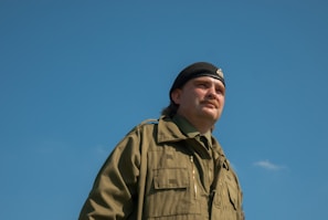 A person wearing a military uniform with a black beret stands against a clear blue sky, looking off into the distance. The uniform is green with several pockets, suggesting practicality and readiness.
