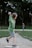 A young boy wearing a green sweatshirt and gray shorts is in mid-action, throwing a baseball. He stands on a dirt mound in a park setting, with lush greenery and a blurred figure in the background.