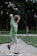 A young boy wearing a green sweatshirt and gray shorts is in mid-action, throwing a baseball. He stands on a dirt mound in a park setting, with lush greenery and a blurred figure in the background.