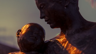 A close-up of a bronze sculpture depicting a mother tenderly holding her child. The lighting casts a warm glow on the figures, highlighting their faces and creating a serene and intimate atmosphere.