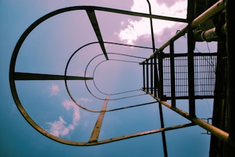 A view from below looking up at a metal ladder enclosed by a circular safety cage. The structure stands against a backdrop of a cloudy sky with sunlight filtering through.
