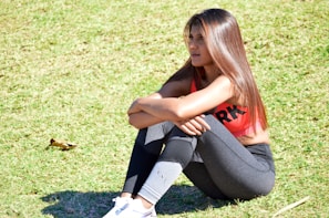 A woman wearing a pink sports bra and black leggings is sitting on grass, resting her arms on her knees with a pensive expression. She has long brown hair and is wearing white sneakers.