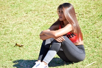 A woman wearing a pink sports bra and black leggings is sitting on grass, resting her arms on her knees with a pensive expression. She has long brown hair and is wearing white sneakers.