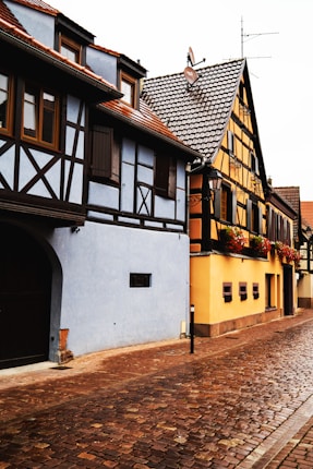 A quaint street lined with half-timbered houses. The buildings feature a mix of blue and yellow exteriors with dark wooden beams, traditional tiled roofs, and intricate details. Flower boxes with vibrant blooms decorate the windows, and a cobblestone path leads between the houses.