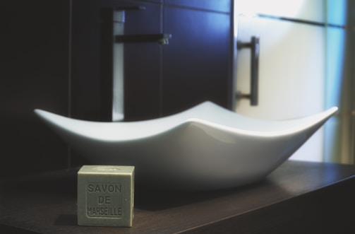 A modern bathroom setting featuring a stylish, white, sculptural sink on a dark countertop. In the foreground is a cube of 'Savon de Marseille' soap, adding a touch of vintage elegance. The background includes dark cabinetry and sleek metal fixtures, contributing to a minimalist aesthetic.