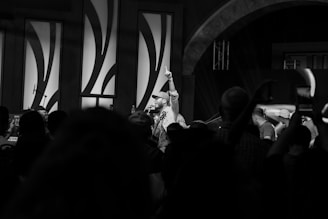 A black and white image captures a lively scene at an indoor event or performance. A central figure holds a microphone while raising a finger in the air, addressing a crowd of people. The audience is engaged, with some capturing the moment on their phones. The background features decorative wall panels and stage lighting.