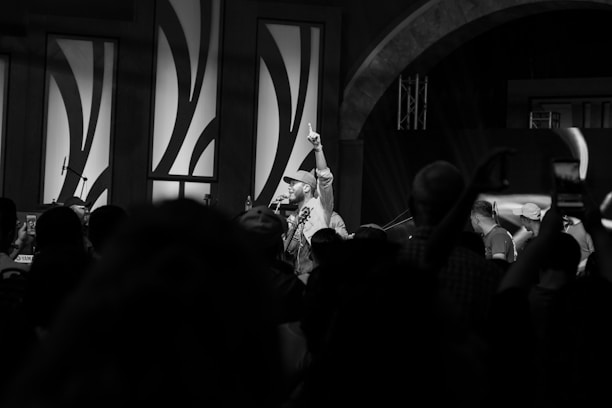 A black and white image captures a lively scene at an indoor event or performance. A central figure holds a microphone while raising a finger in the air, addressing a crowd of people. The audience is engaged, with some capturing the moment on their phones. The background features decorative wall panels and stage lighting.