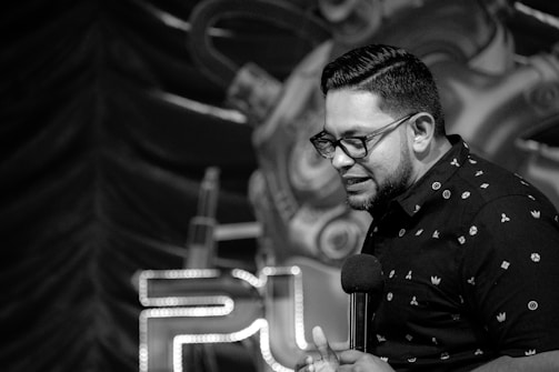 A person holds a microphone, standing in front of a distinctive backdrop that includes stylized mechanical elements and illuminated letters. The scene is captured in black and white, giving it a classic, timeless feel.